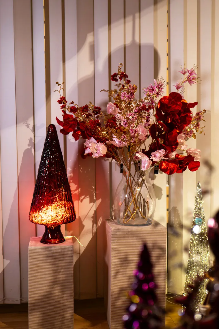 Albero di vetro bordeaux illuminato accanto a un vaso di fiori rossi e rosa